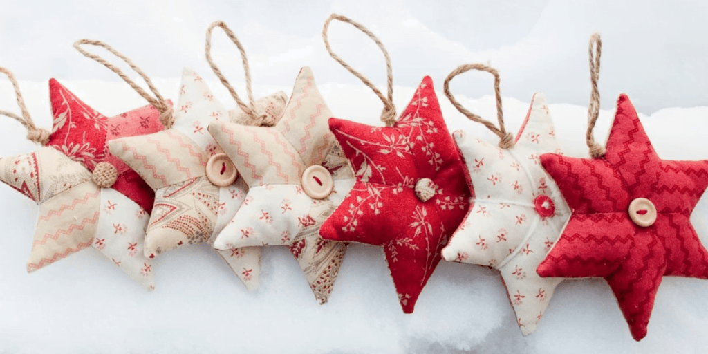 A row of eight star-shaped fabric ornaments in various red and white patterns hang by jute strings. These charming stars, each with a button at the center, are displayed against a snowy background – a perfect Christmas idea for festive decor.