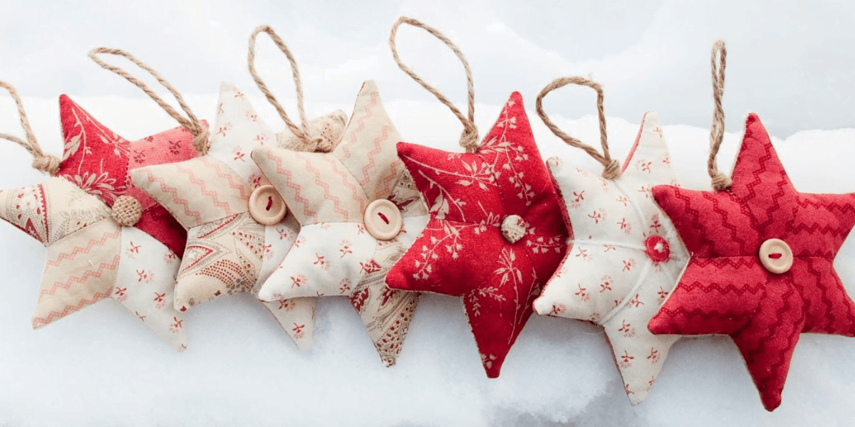 A row of eight star-shaped fabric ornaments in various red and white patterns hang by jute strings. These charming stars, each with a button at the center, are displayed against a snowy background – a perfect Christmas idea for festive decor.